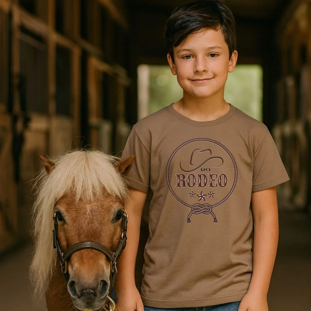 A young boy wearing a brown graphic t-shirt stands next to a small pony in a rustic barn setting. The t-shirt features a dark brown circular graphic that reads 'LIFE RODEO' with a cowboy hat and rope design.