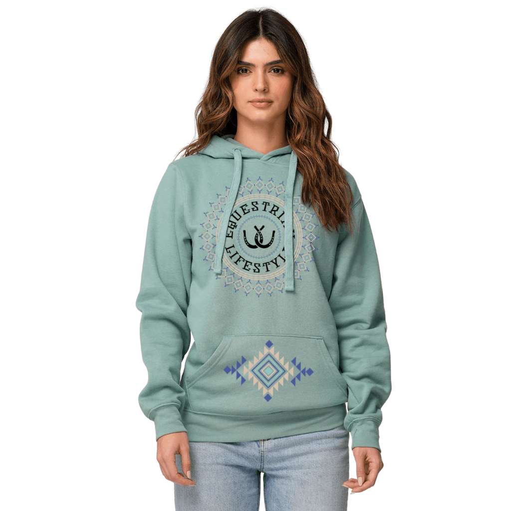 A woman modeling a sage green pullover hoodie with a circular equestrian Lifestyle' graphic on the chest and a blue and cream geometric diamond pattern on the front pocket, set against a plain white background.