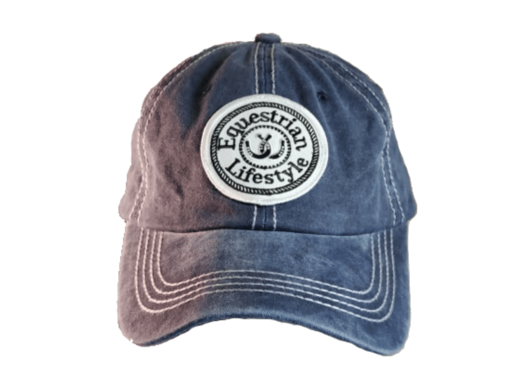 A washed blue denim baseball cap with a circular white embroidered patch on the front that reads 'Equestrian Lifestyle' with a horseshoe symbol. The cap features a faded color gradient transitioning from a dusty mauve-pink on the left to a medium blue on the right, with white contrast stitching.