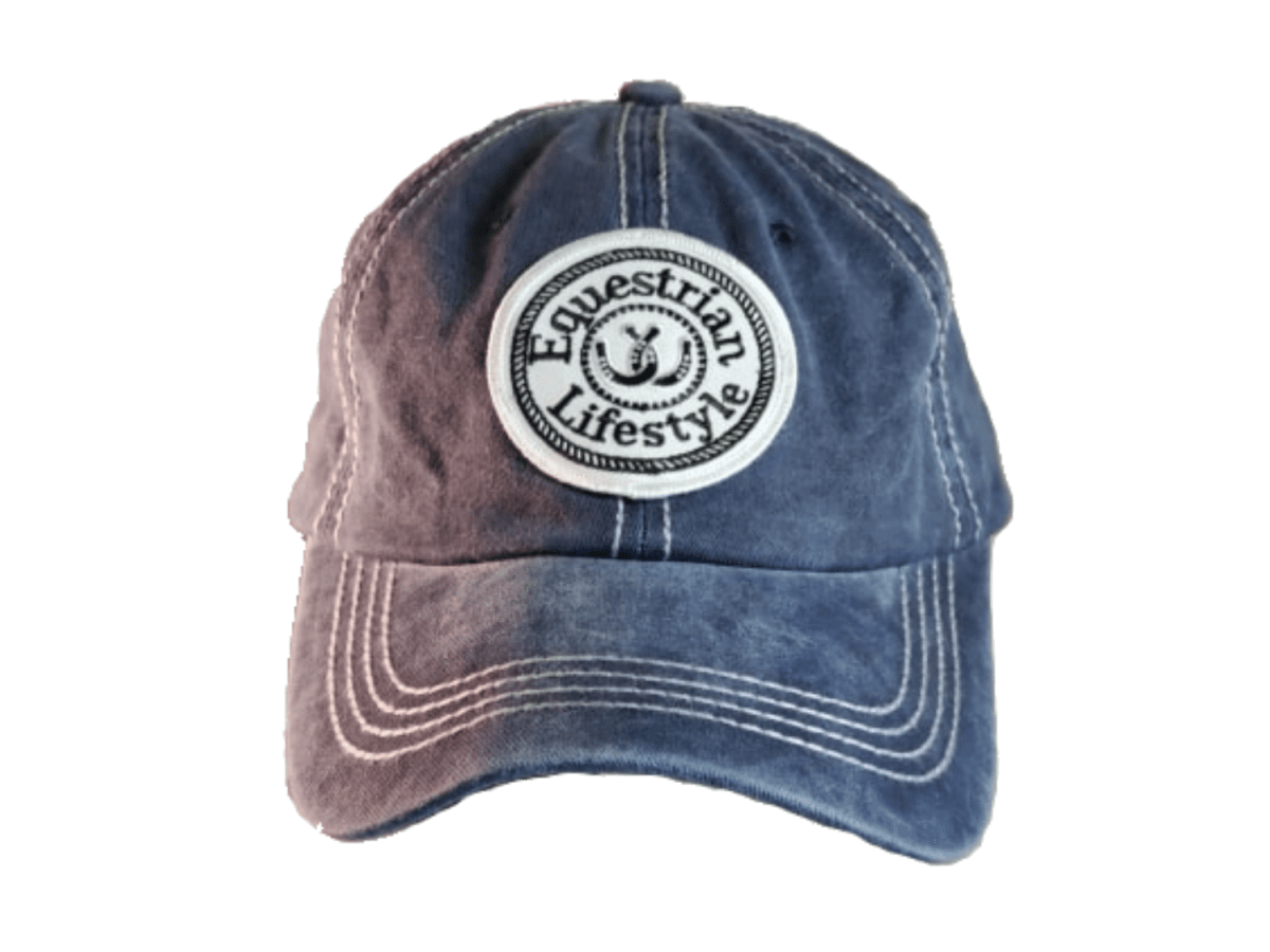 A washed blue denim baseball cap with a circular white embroidered patch on the front that reads 'Equestrian Lifestyle' with a horseshoe symbol. The cap features a faded color gradient transitioning from a dusty mauve-pink on the left to a medium blue on the right, with white contrast stitching.