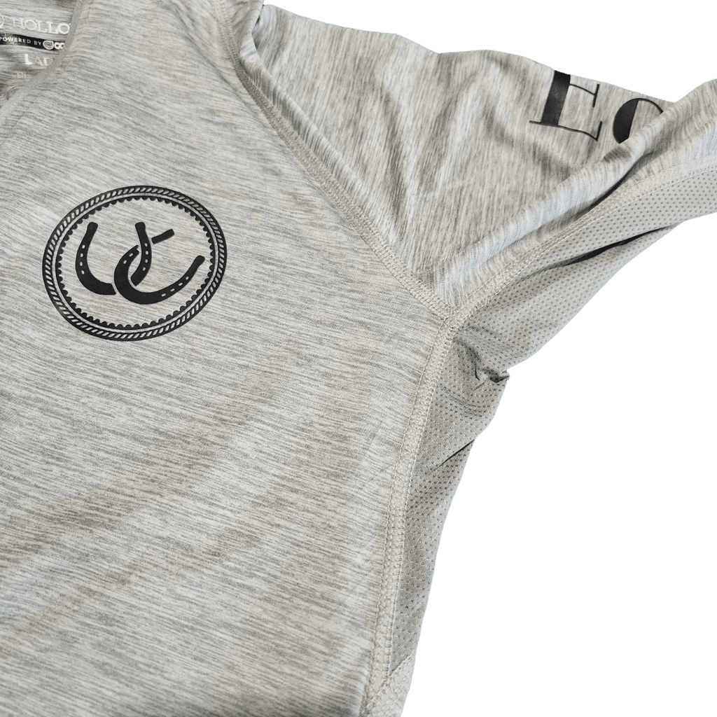Close-up of a light heather gray athletic top showing the chest area with a black circular horseshoe logo and part of a black printed wordmark on the shoulder.