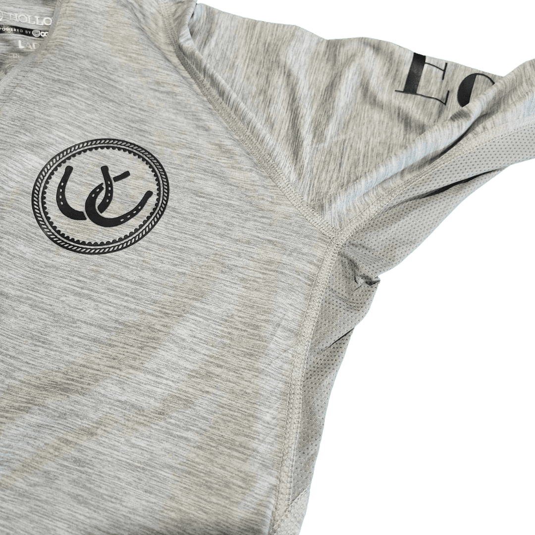 Close-up of a light heather gray athletic top showing the chest area with a black circular horseshoe logo and part of a black printed wordmark on the shoulder.