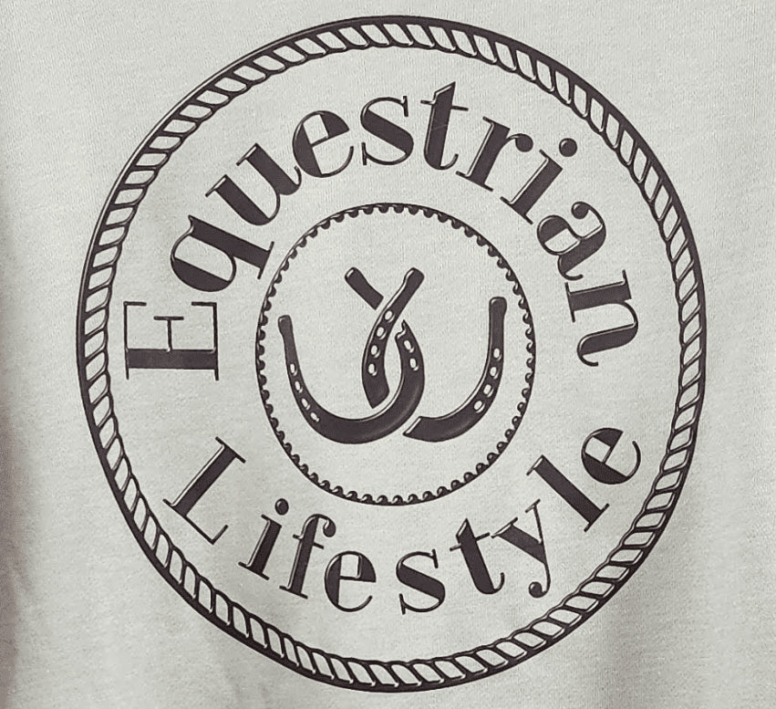 A close-up of a light sage fabric surface featuring a circular black graphic print with the words 'Equestrian Lifestyle' and a stylized horseshoe design in the center.