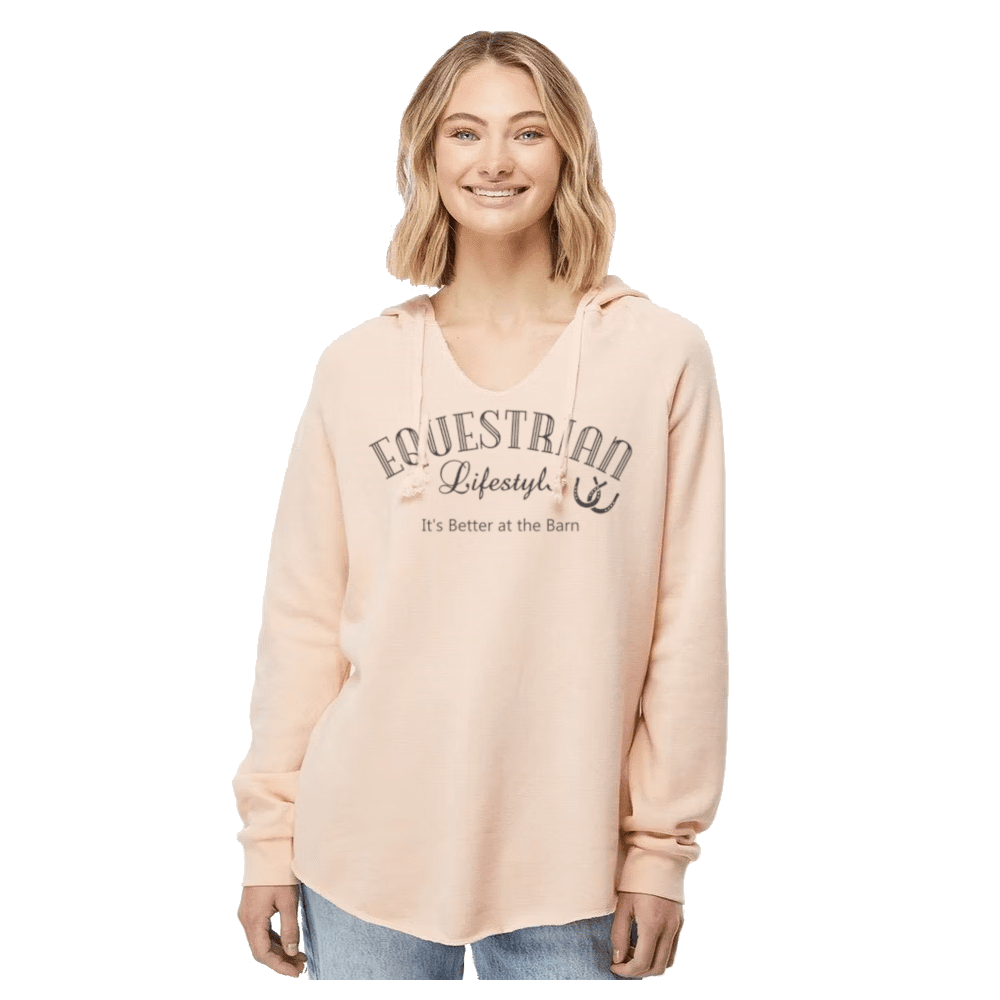A woman wearing a light peach-pink pullover hoodie with 'EQUESTRIAN Lifestyle' and 'It's Better at the Barn' printed on the front, paired with blue jeans, against a plain white background.