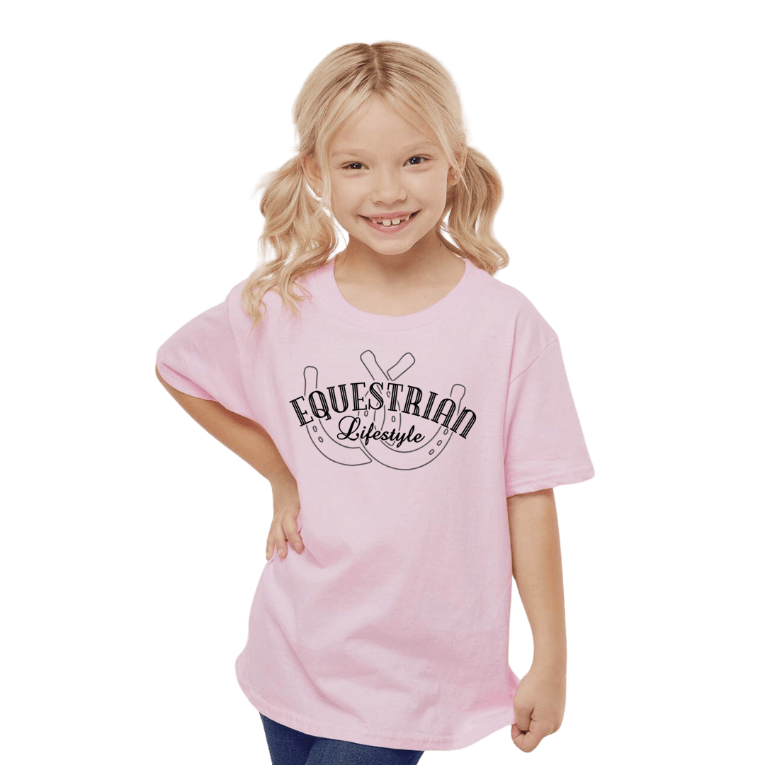 A young blonde girl with pigtails smiling while wearing a light pink t-shirt with a black graphic that reads 'EQUESTRIAN Lifestyle' over  a horseshoe. She is standing against a solid white background.