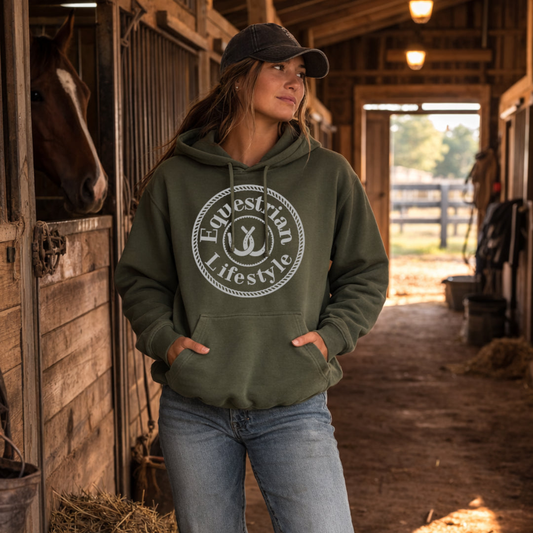 A woman wearing an olive green hoodie with a white circular 'Equestrian Lifestyle' graphic stands in a barn aisle with a horse visible in a stall on the left.