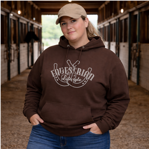A woman wearing a brown hoodie with a white 'Equestrian Lifestyle' graphic stands in a horse stable corridor, wearing a beige baseball cap and blue jeans.