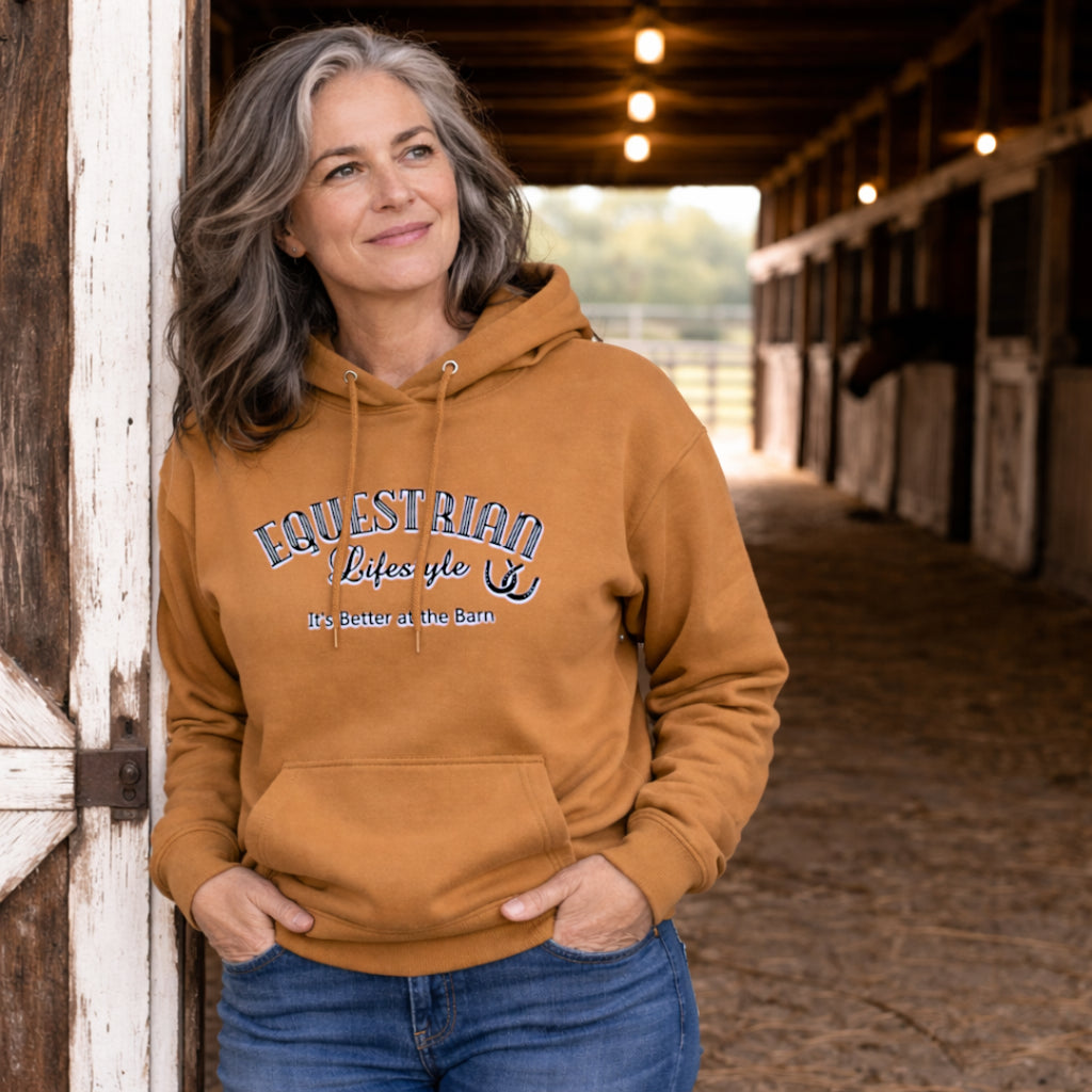 A woman with gray hair stands in a barn doorway wearing a camel brown hoodie with 'Equestrian Lifestyle'  and its better at the barn printed on the chest, paired with blue jeans.