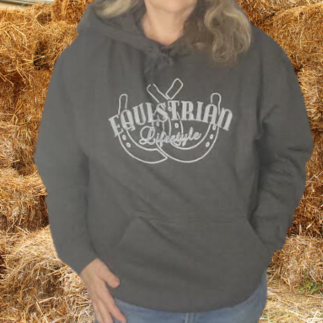 A person wearing a dark charcoal gray hoodie with a white graphic that reads 'EQUESTRIAN Lifestyle' over  horseshoes, standing in front of a backdrop of hay bales.
