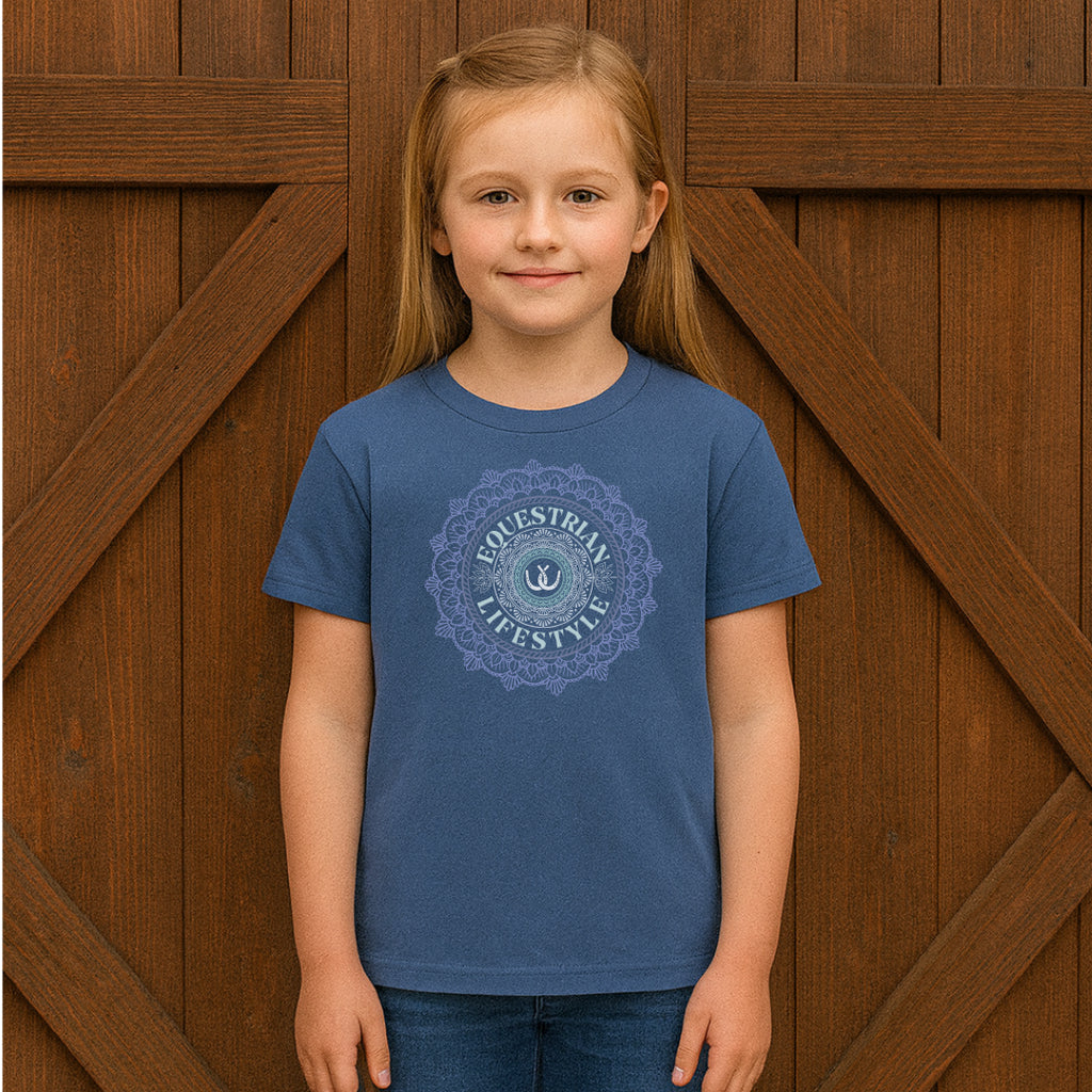 A young girl with light brown hair stands in front of a rustic wooden barn door, wearing a slate blue crewneck t-shirt with a light blue circular mandala graphic that reads 'equestrianLIFESTYLE'.