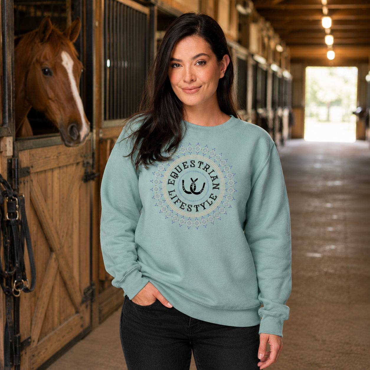 A woman standing in a horse stable wearing a sage green crewneck sweatshirt with a black and white 'Equestrian Lifestyle' graphic on the chest. A horse is visible in the stall to her left.