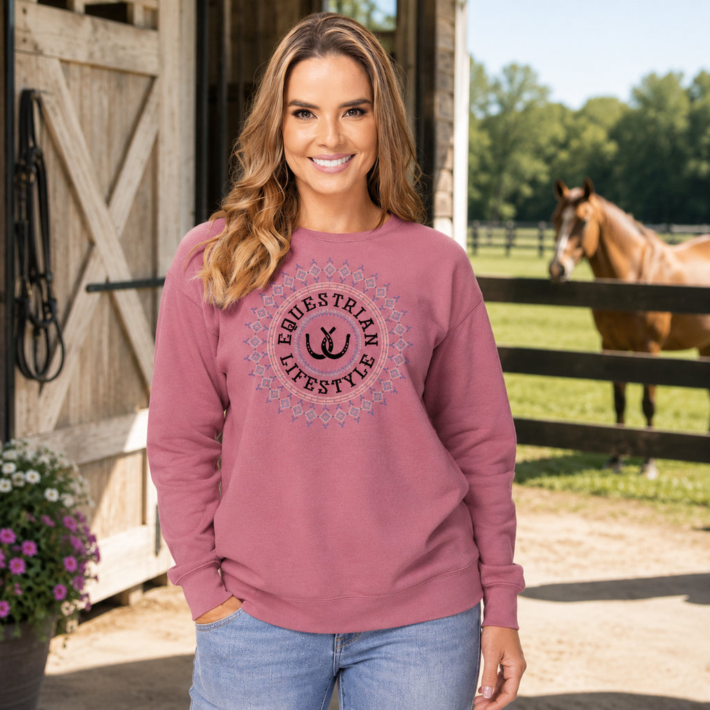 A woman stands outdoors in front of a wooden barn and fence, wearing a dusty rose crewneck sweatshirt with a circular 'Equestrian Lifestyle' graphic on the chest, paired with light blue jeans. A brown horse is visible in the background.