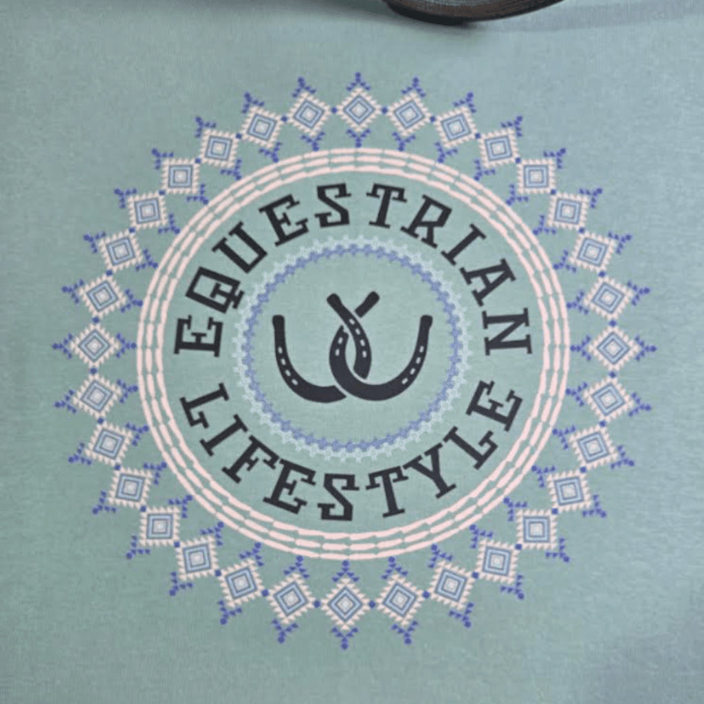 A close-up of a light teal t-shirt featuring a circular graphic with the words 'EQUESTRIAN LIFESTYLE' in dark gray lettering, surrounding a central double-horseshoe design, all framed by a geometric diamond aztec pattern.