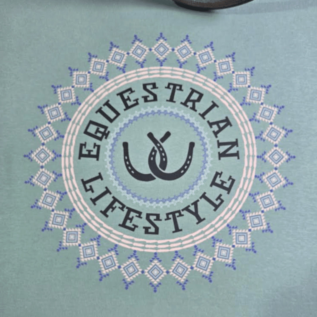 A close-up of a light teal t-shirt featuring a circular graphic with the words 'EQUESTRIAN LIFESTYLE' in dark gray lettering, surrounding a central double-horseshoe design, all framed by a geometric diamond aztec pattern.