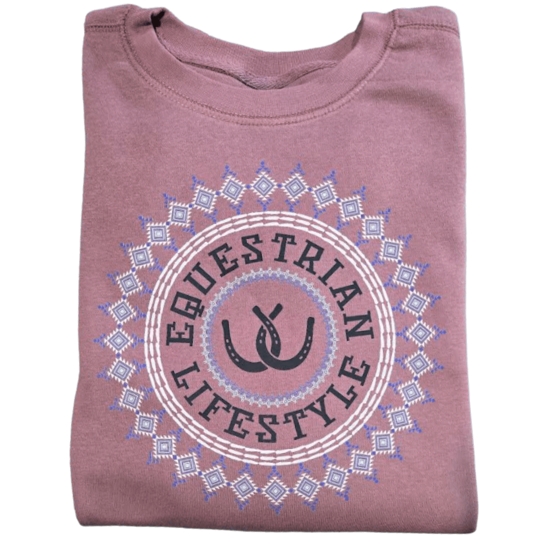 A folded dusty rose t-shirt featuring a circular graphic with the words 'EQUESTRIAN LIFESTYLE' in black, flanked by two black horseshoes, all enclosed in a white and light blue geometric aztec pattern.