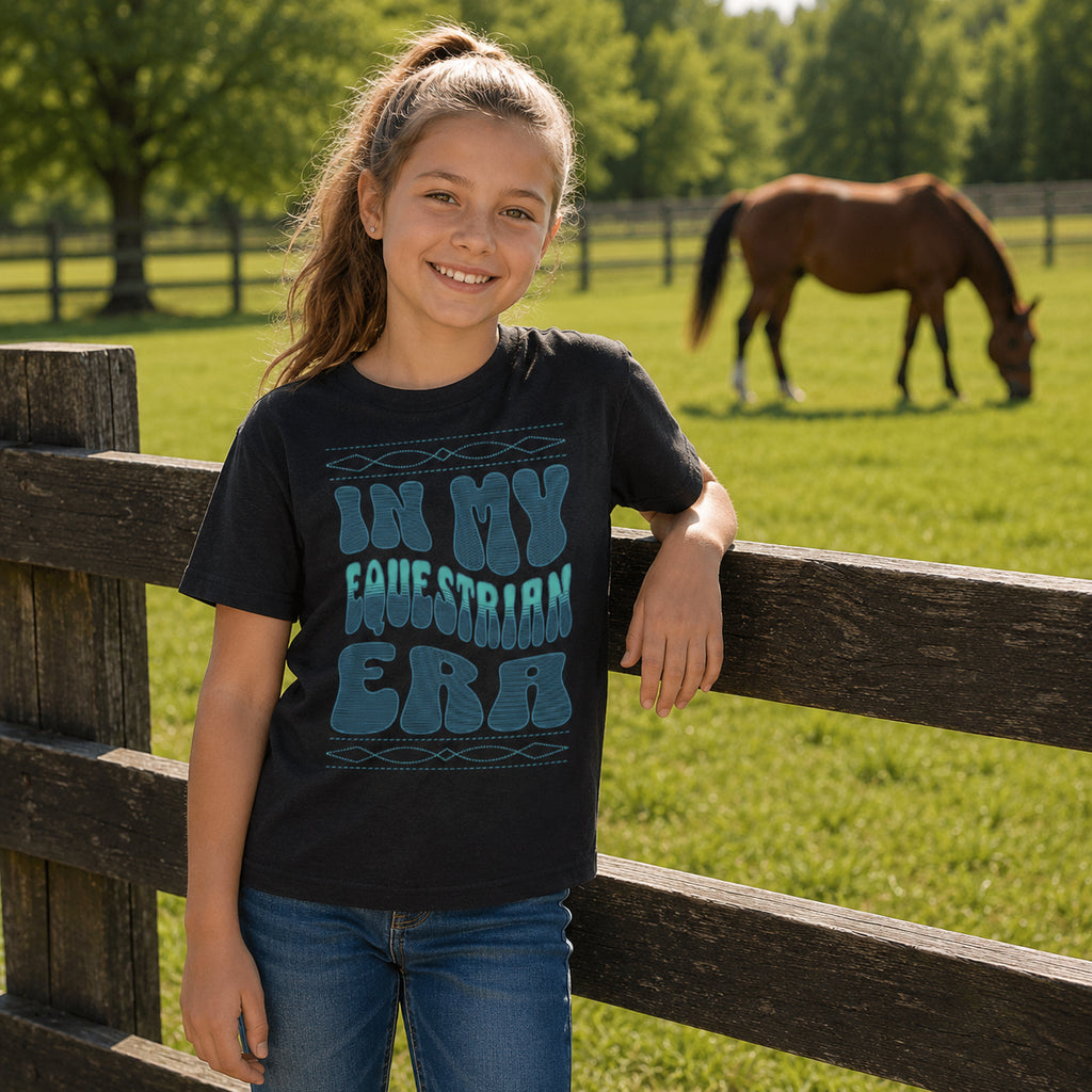 A young girl smiling and leaning on a wooden fence in a grassy paddock, wearing a black graphic t-shirt that reads 'IN MY EQUESTRIAN ERA' in teal lettering. A brown horse is grazing in the background.
