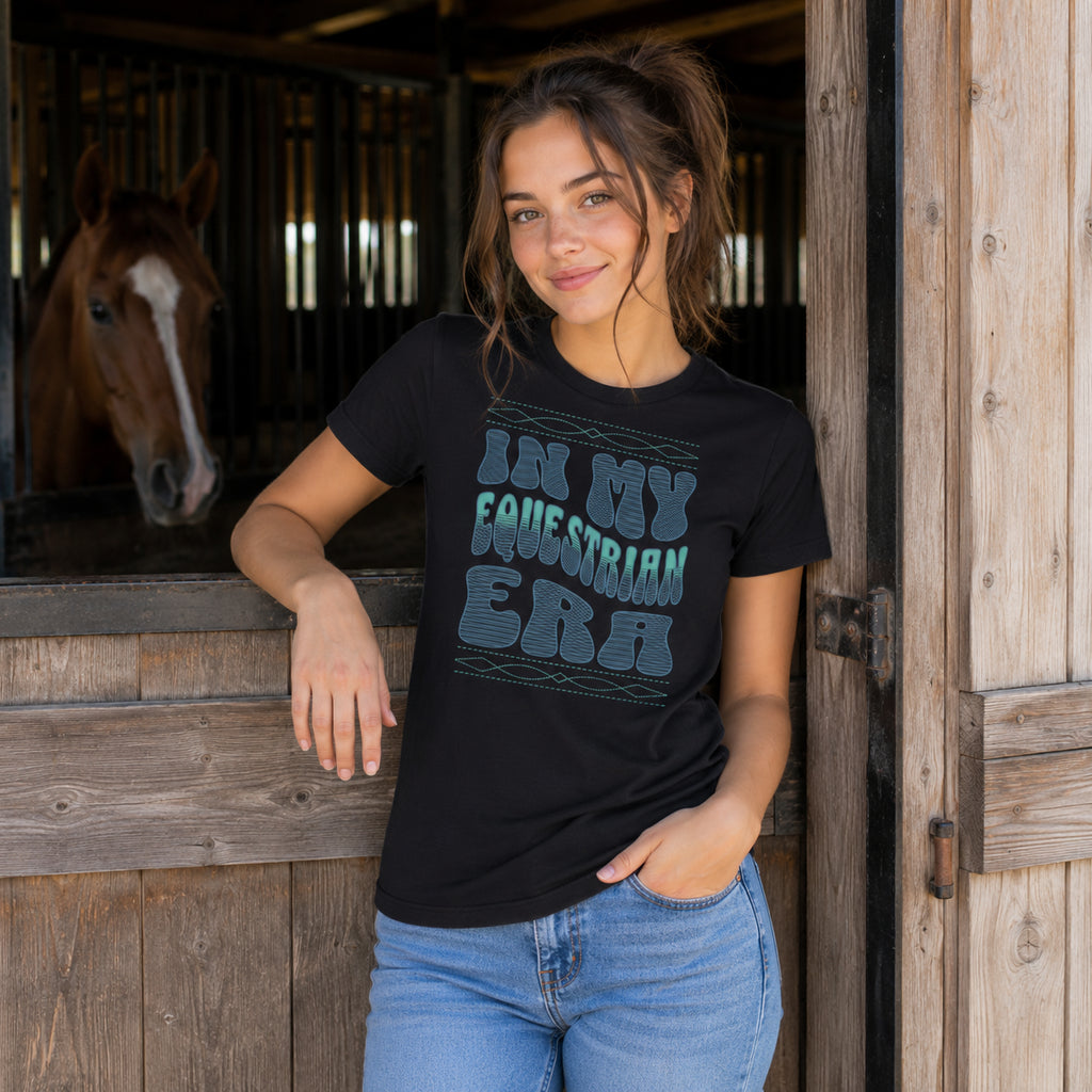 A woman wearing a black graphic t-shirt with the text 'IN MY EQUESTRIAN ERA' in a distressed teal font, posing in front of a rustic wooden stable door with a horse visible in the background.