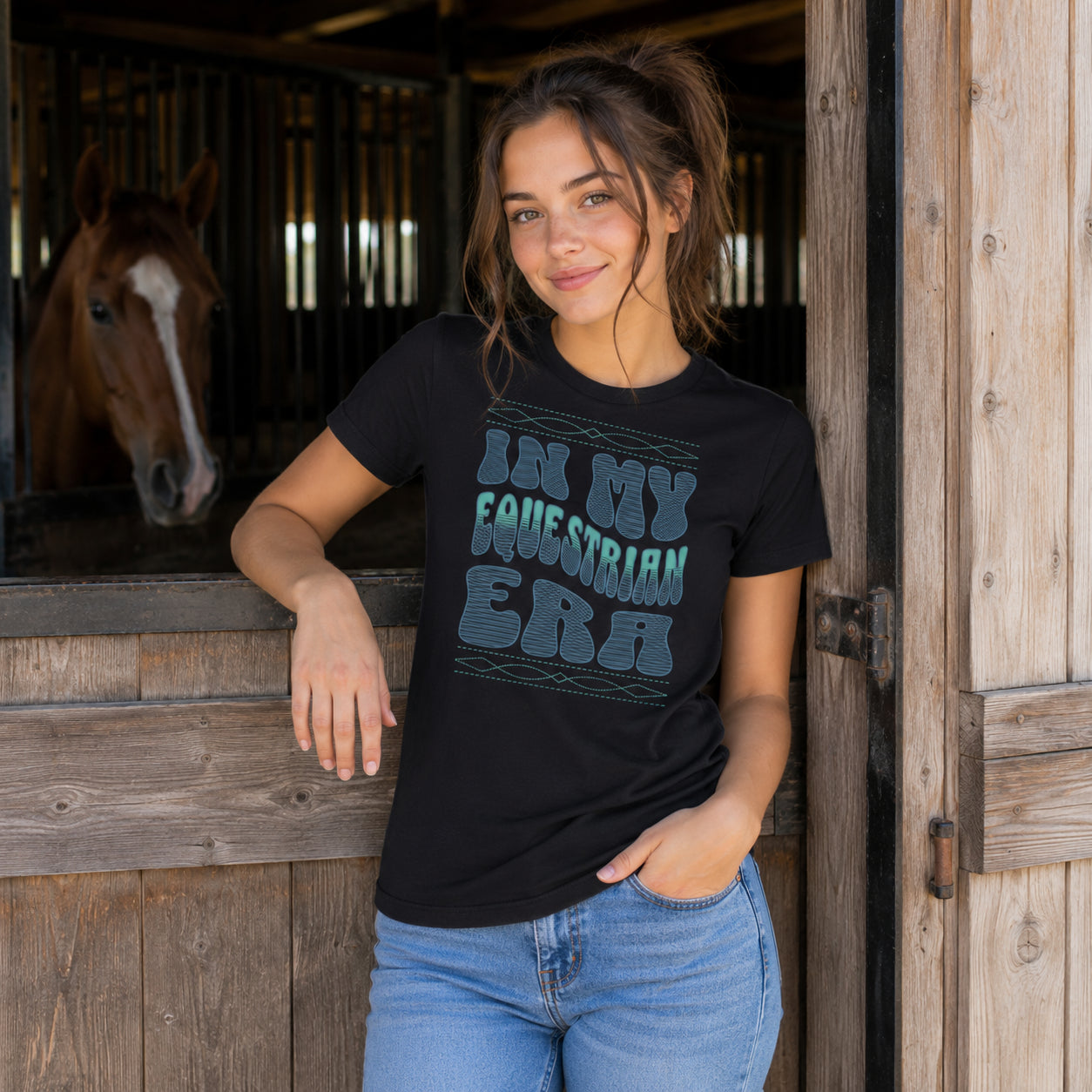 A woman wearing a black graphic t-shirt with the text 'IN MY EQUESTRIAN ERA' in a distressed teal font, posing in front of a rustic wooden stable door with a horse visible in the background.