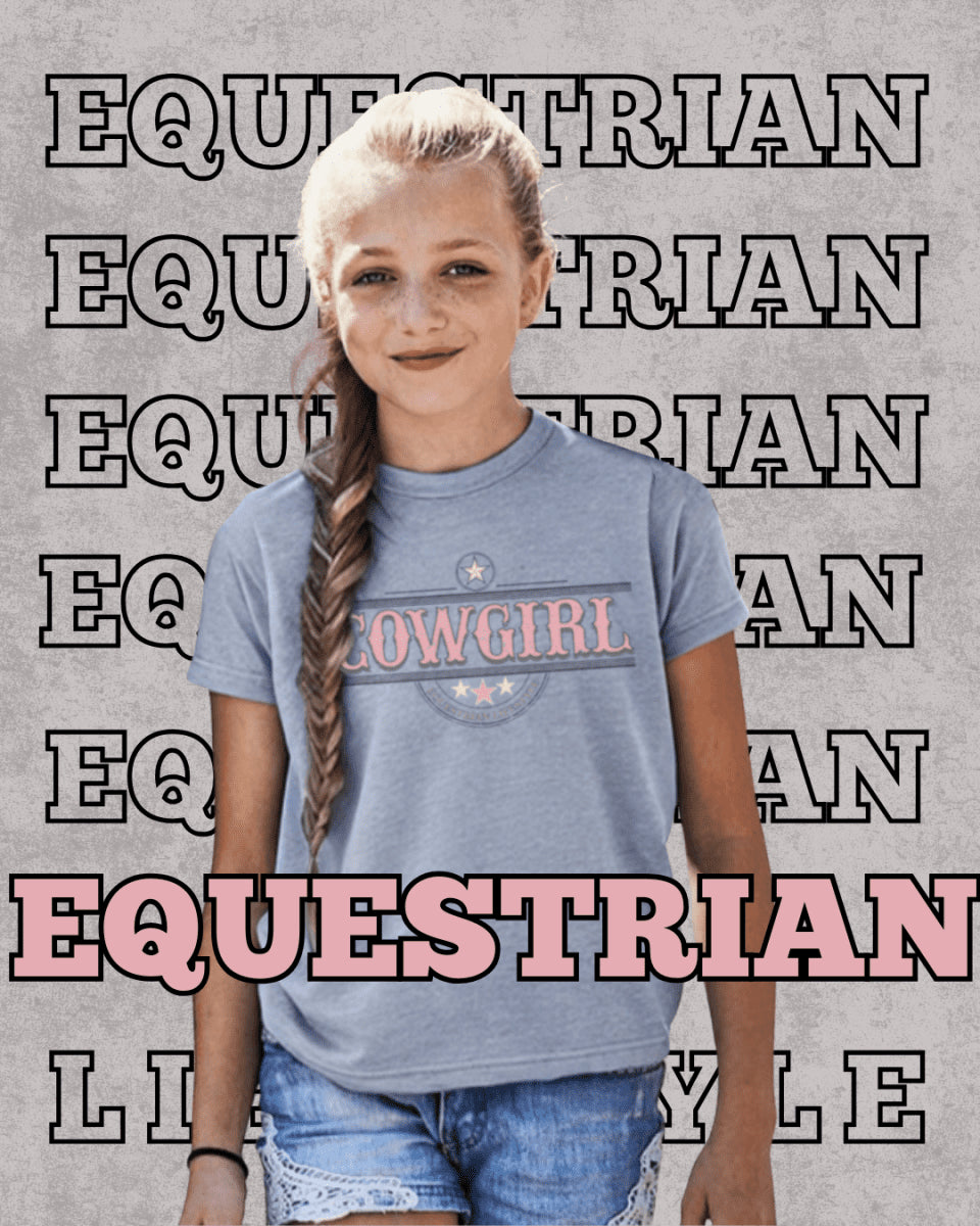 A young girl with blonde hair in a braid wears a heathered blue t-shirt featuring a 'COWGIRL' graphic with the word 'EQUESTRIAN' in pink. The background is a textured gray wall with repeated 'EQUESTRIAN' text outlines, and a large 'EQUESTRIAN' text overlay at the bottom.