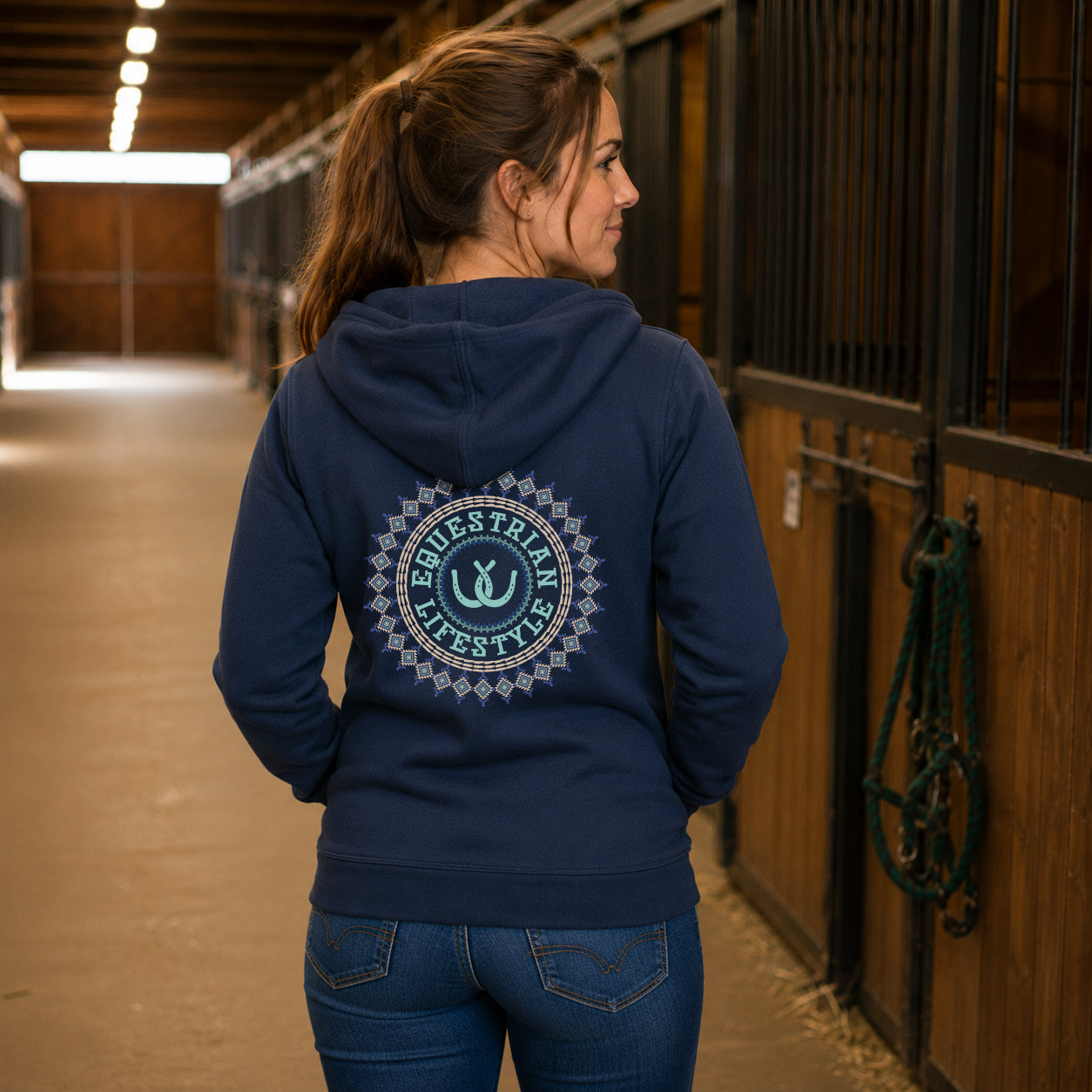 A woman standing in a horse stable wearing a navy blue hoodie with a large circular 'Equestrian Lifestyle' logo printed on the back, paired with blue jeans.