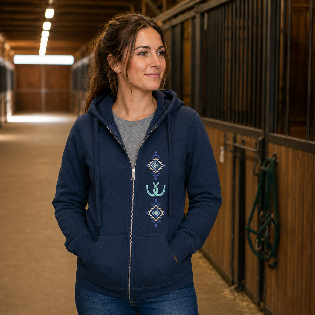 A woman wearing a navy blue zip-up hoodie with a gray crewneck shirt and blue jeans, standing in a horse stable corridor.
