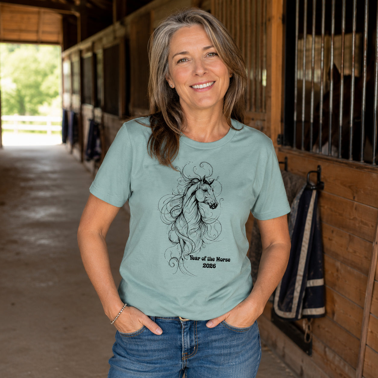 A smiling woman stands in a horse stable wearing a sage green graphic t-shirt with a black line art illustration of a horse and the text 'Year of the Horse 2026'.