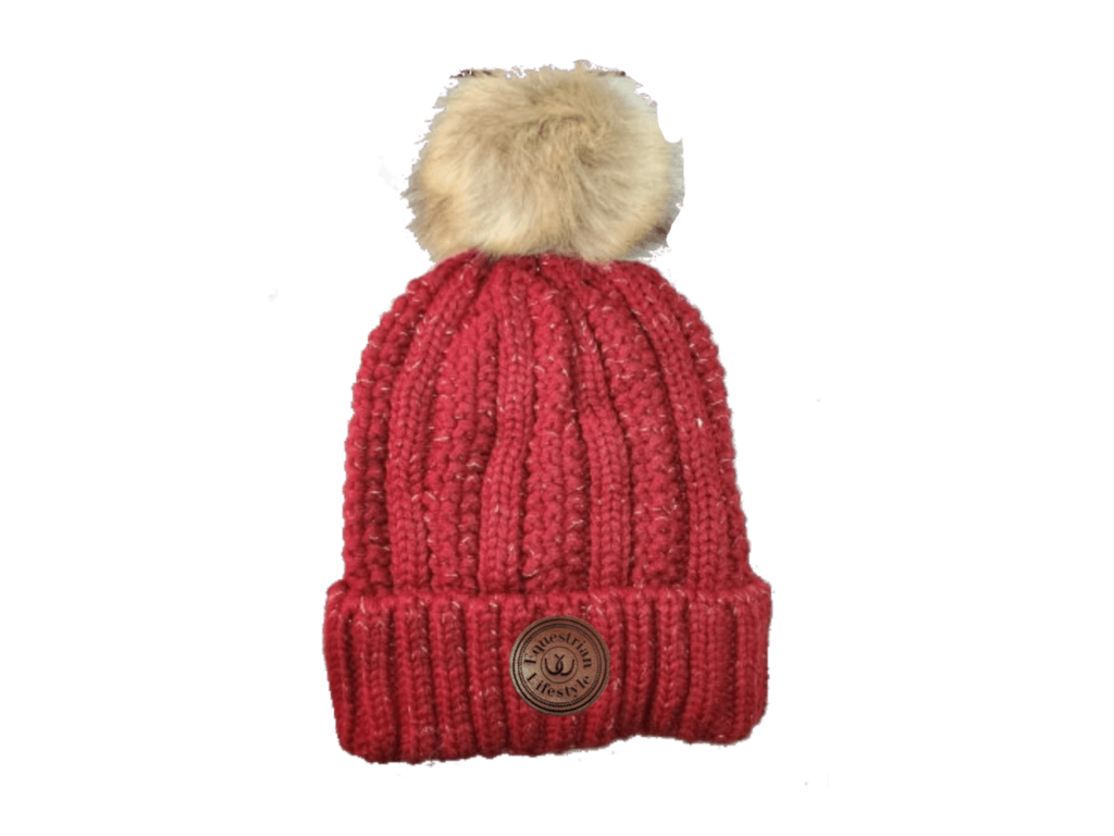 A red ribbed knit beanie hat with a fluffy beige faux-fur pom-pom on top and a circular brown equestrin lifestyle brand patch on the front cuff, shown against a plain white background.