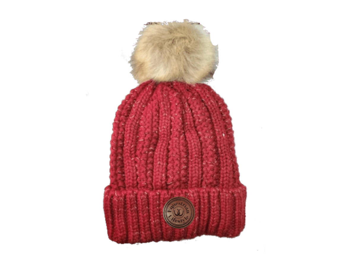 A red ribbed knit beanie hat with a fluffy beige faux-fur pom-pom on top and a circular brown equestrin lifestyle brand patch on the front cuff, shown against a plain white background.