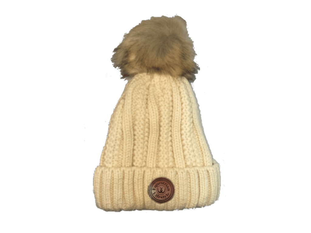 A cream-colored cable-knit beanie hat with a brown faux-fur pom-pom on top and a circular brown leather equestrian lifestyle patch on the front cuff.
