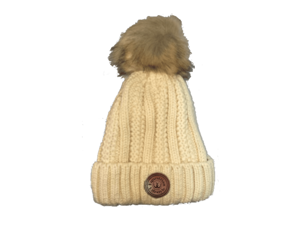 A cream-colored cable-knit beanie hat with a brown faux-fur pom-pom on top and a circular brown leather equestrian lifestyle patch on the front cuff.