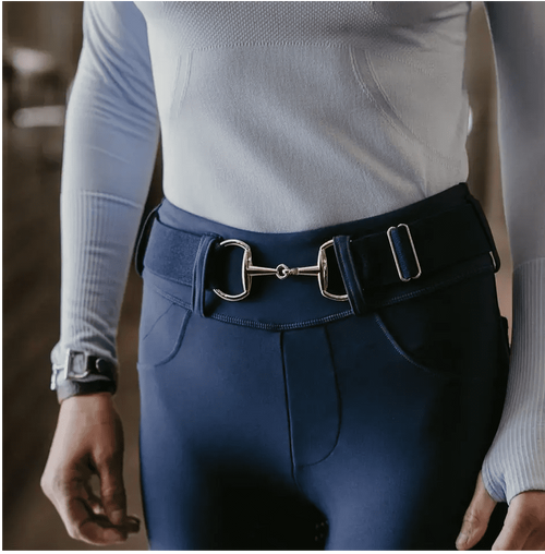 A close-up of a person wearing navy blue equestrian riding pants with a matching navy belt featuring a silver horsebit buckle, paired with a light blue ribbed long-sleeve top.