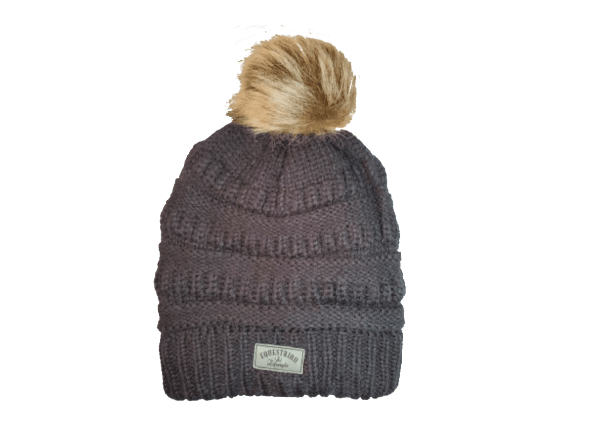 A dark gray ribbed knit beanie hat with a fluffy tan faux-fur pom-pom on top and a small white brand patch on the front cuff. The brand name 'equestrian lifestyle is visible on the patch.