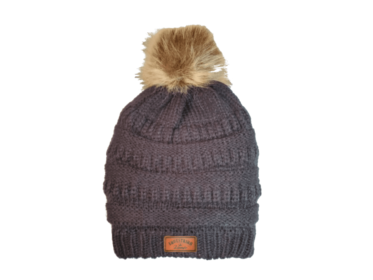 A dark gray ribbed knit beanie hat with a fluffy tan faux-fur pom-pom on top and a small brown leatherette equestrian lifestyle  brand patch on the front cuff.