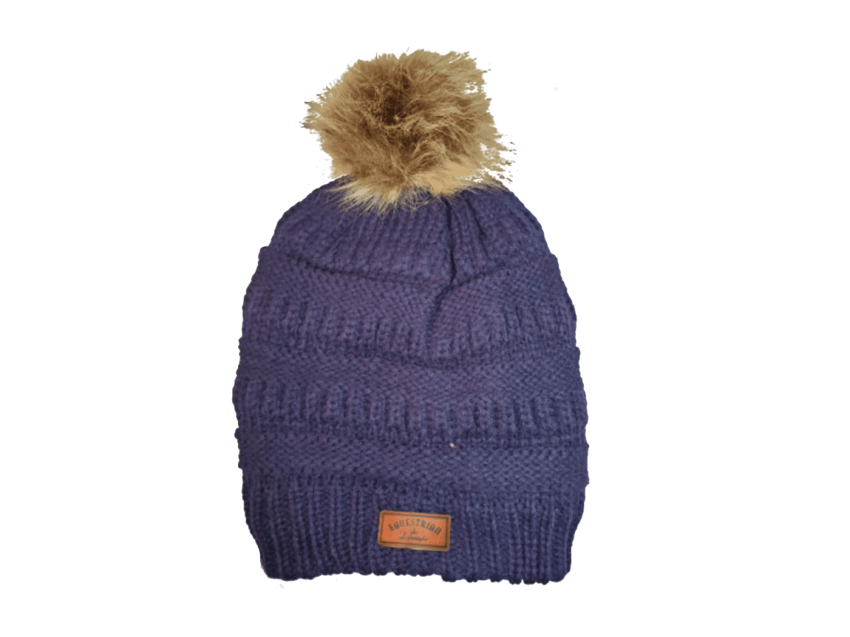 A ribbed knit beanie hat in a dark purple color, featuring a fluffy tan faux-fur pom-pom on top and a small brown leatherette equestrian lifestyle brand patch near the bottom cuff.