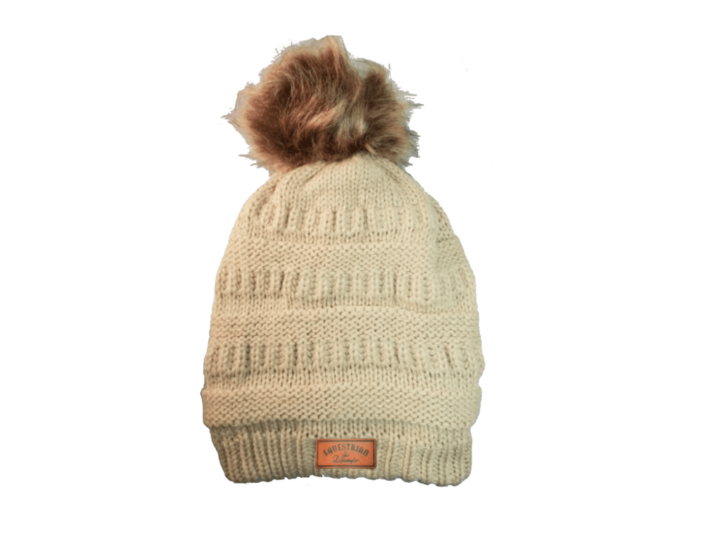 A cream-colored ribbed knit beanie hat with a brown faux fur pom-pom on top and a small brown leatherette equestrian lifestyle brand patch near the brim.