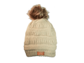 A cream-colored ribbed knit beanie hat with a brown faux fur pom-pom on top and a small brown leatherette equestrian lifestyle brand patch near the brim.