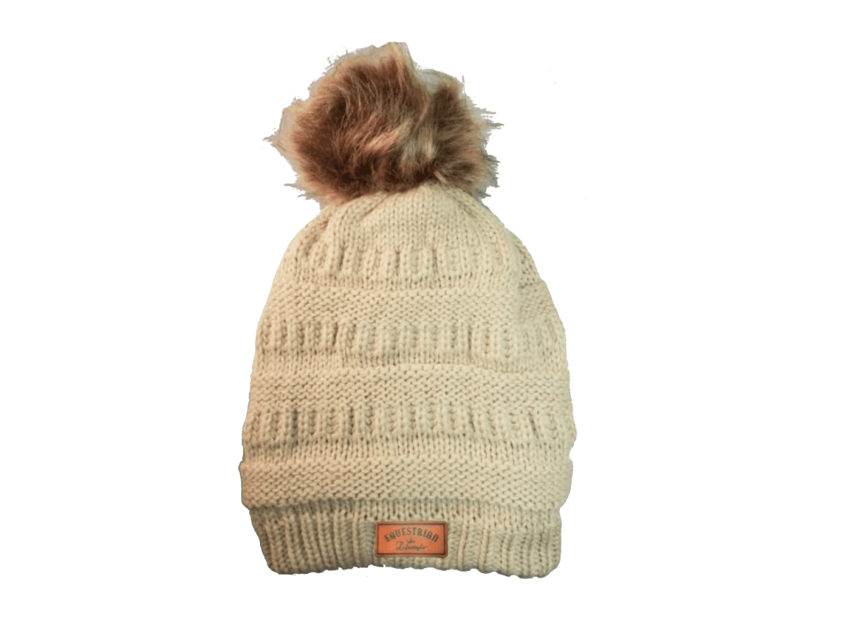 A cream-colored ribbed knit beanie hat with a brown faux fur pom-pom on top and a small brown leatherette equestrian lifestyle brand patch near the brim.