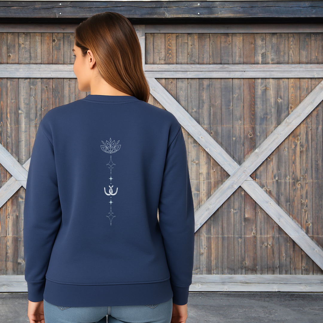 A woman viewed from the back wearing a navy blue crewneck sweatshirt with a white lotus and horseshoe graphic on the back, standing in front of a rustic wooden barn door.