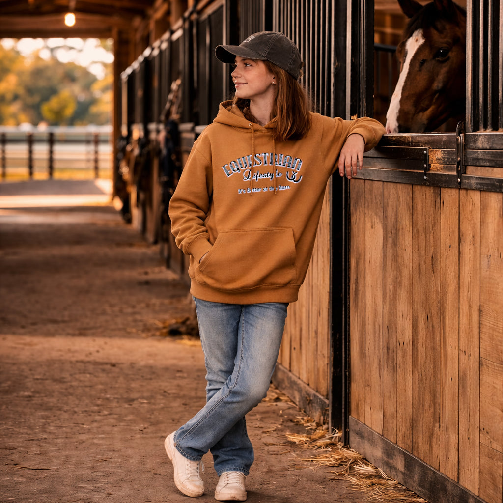 A woman stands in a barn aisle wearing a camel-colored hoodie with 'equestrian lifestyle' printed on the front, paired with blue jeans and a dark baseball cap. She is leaning against a wooden stable door next to a horse.
